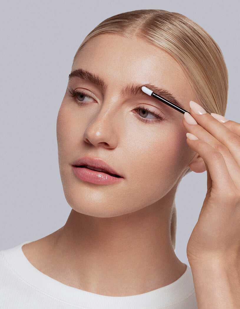 Woman applying brow serum to her eyebrows with a applicator against a neutral background